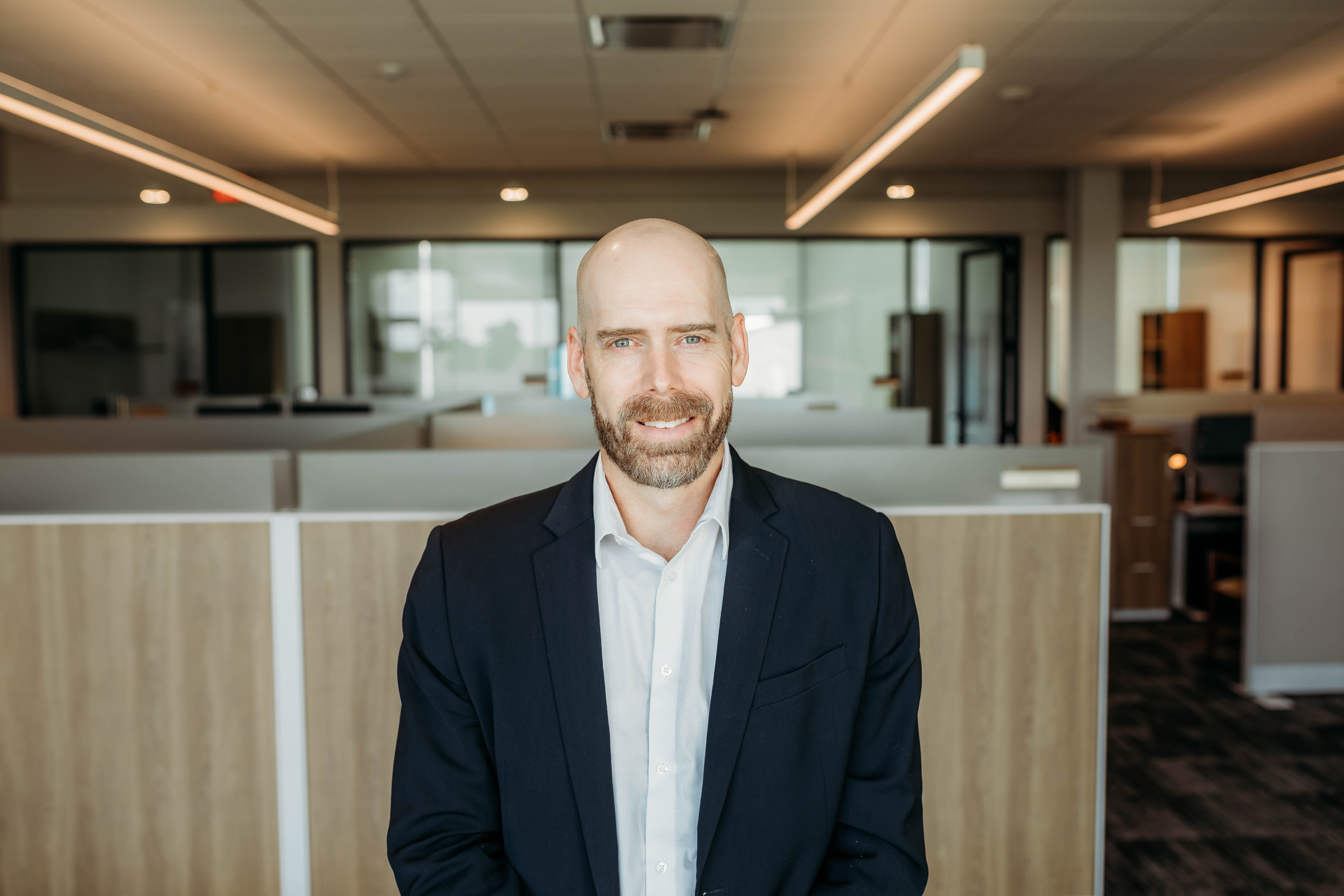 Man with shaved head and beard wearing a navy suit jacket and white button stands in front of interior offices