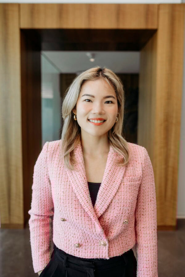Young woman with blonde hair standing in front of a hallway wearing a pink blazer