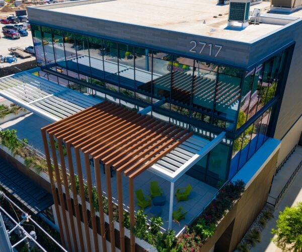 Aerial exterior photo of a modern office building showing grey paneling, lots of windows and wood geometric accents over a balcony with trees surrounding